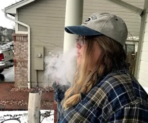 Long haired person wearing a hat standing outside smoking